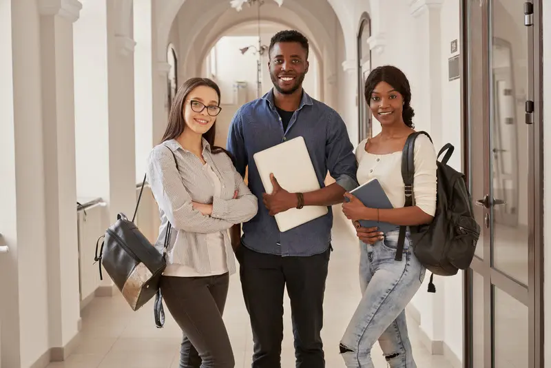 três estudantes com mochilas, um segurando um laptop e outro um caderno, em um corredor com arcadas
