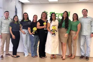 Grupo de pessoas em pé em escritório com logo do Sicredi ao fundo; três pessoas seguram mascotes amarelos em forma de abelha