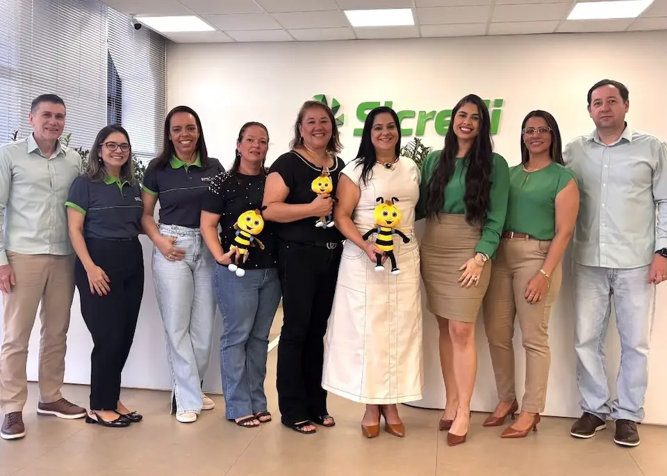 Grupo de pessoas em pé em escritório com logo do Sicredi ao fundo; três pessoas seguram mascotes amarelos em forma de abelha