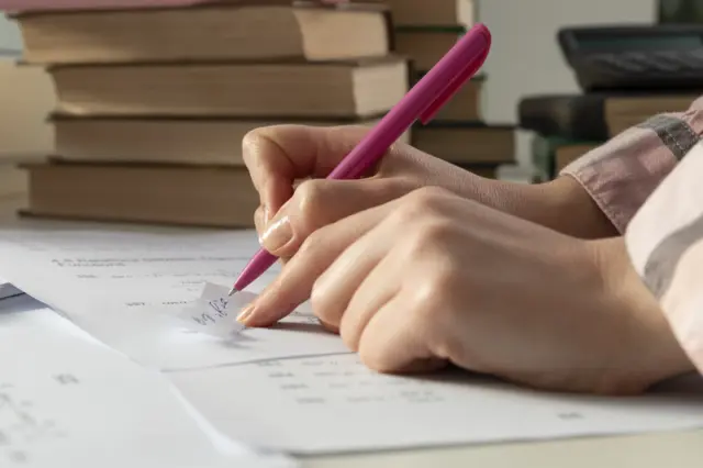 Mãos de pessoa escrevendo com caneta rosa sobre folhas de papel, com pilha de livros desfocada ao fundo