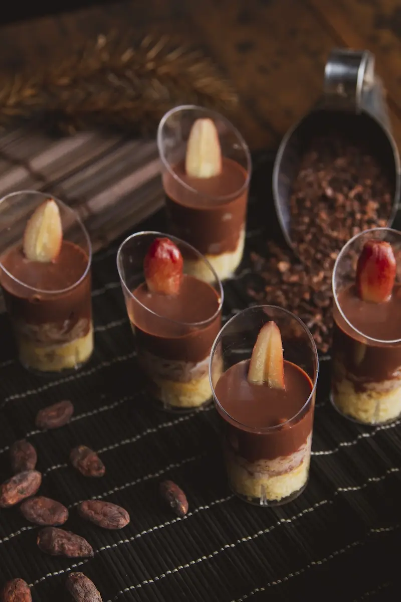 pequenos copos plásticos com camadas de creme e cobertura de chocolate, cada um guarnecido com uma fatia de fruta, sobre tecido escuro com grãos de cacau ao lado