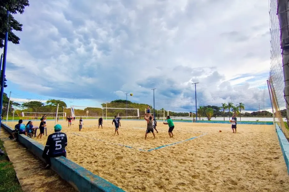 Jogadores praticando vôlei em quadra de areia com espectadores sentados na mureta e céu nublado