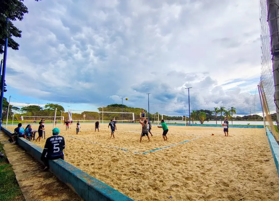 Jogadores praticando vôlei em quadra de areia com espectadores sentados na mureta e céu nublado