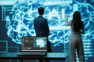 Notebook em mesa com visualização de cérebro digital na tela e duas pessoas em pé diante de um grande painel com padrões neurais e a sigla 'AI'