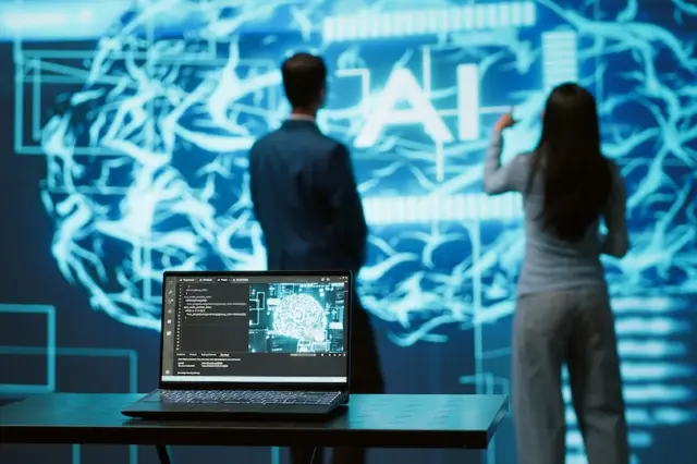 Notebook em mesa com visualização de cérebro digital na tela e duas pessoas em pé diante de um grande painel com padrões neurais e a sigla 'AI'