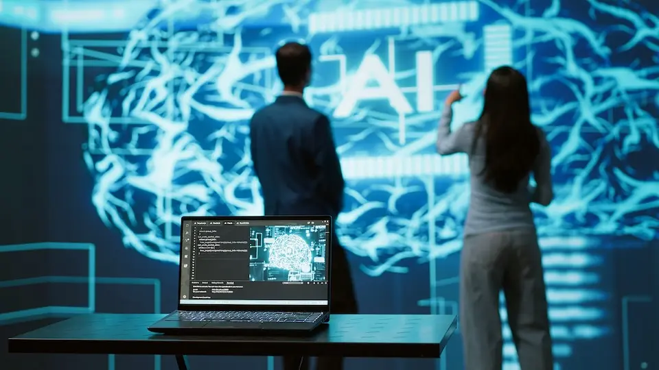 Notebook em mesa com visualização de cérebro digital na tela e duas pessoas em pé diante de um grande painel com padrões neurais e a sigla 'AI'