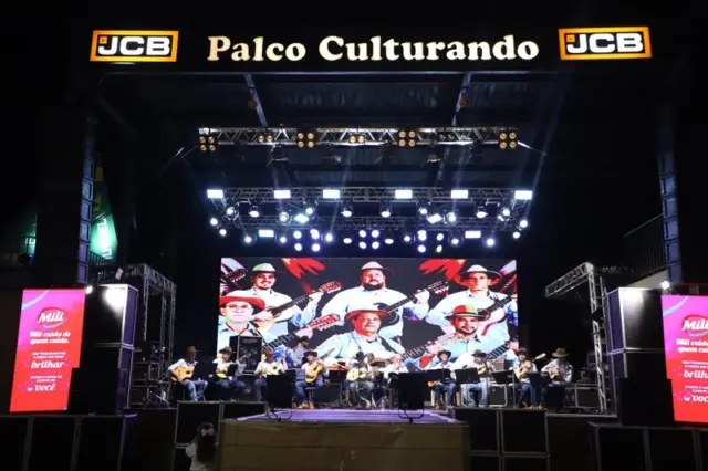 Palco Culturando iluminado com um grupo de músicos tocando violões e outros instrumentos, com telão ao fundo mostrando imagens dos artistas
