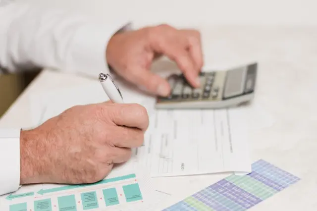 Mãos segurando caneta sobre documentos, com outra mão usando calculadora e planilha colorida ao lado