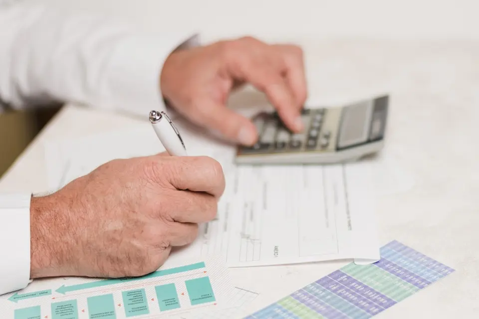Mãos segurando caneta sobre documentos, com outra mão usando calculadora e planilha colorida ao lado