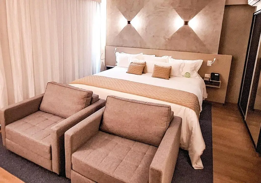 Quarto de hotel com cama king-size, duas poltronas em primeiro plano e cortinas claras