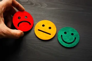 Mão segurando um emoji vermelho triste ao lado de emojis amarelo neutro e verde sorrindo sobre superfície escura