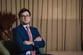 Homem adulto sentado em poltrona, vestindo terno azul, camisa branca e gravata rosa, de óculos, com braços cruzados e cortina bege ao fundo