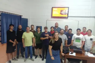 Grupo de cerca de 15 pessoas posando em sala de aula; maioria homens e uma mulher sorrindo à frente, quadro branco e televisão ao fundo