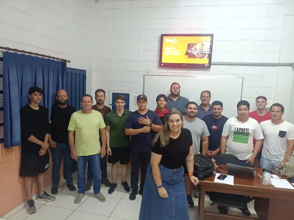 Grupo de cerca de 15 pessoas posando em sala de aula; maioria homens e uma mulher sorrindo à frente, quadro branco e televisão ao fundo