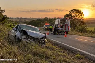 Carro com a frente amassada na margem de uma rodovia, com equipe de resgate, ambulância e cones na pista ao fundo, céu ao pôr do sol e selo 'imagem ilustrativa' no canto