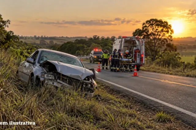 Carro com a frente amassada na margem de uma rodovia, com equipe de resgate, ambulância e cones na pista ao fundo, céu ao pôr do sol e selo 'imagem ilustrativa' no canto