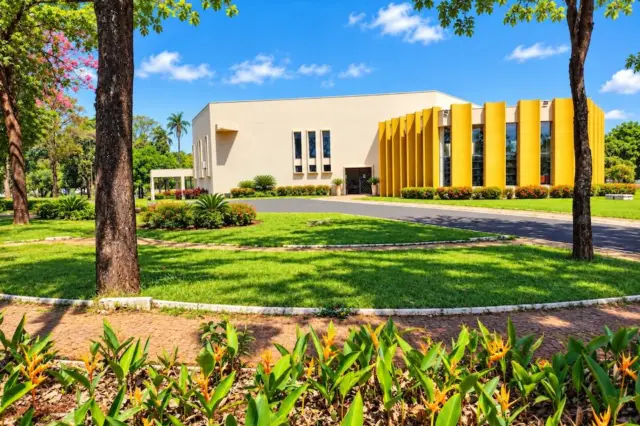 Prédio com fachada branca e painéis verticais amarelos, jardim e canteiros floridos à frente