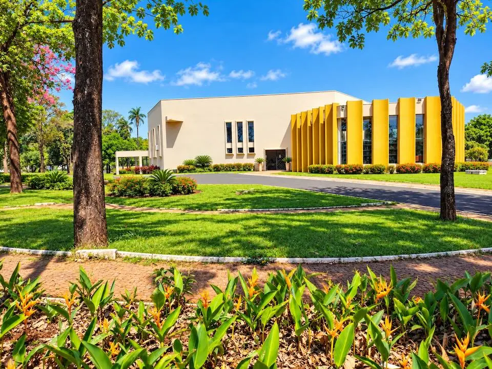 Prédio com fachada branca e painéis verticais amarelos, jardim e canteiros floridos à frente
