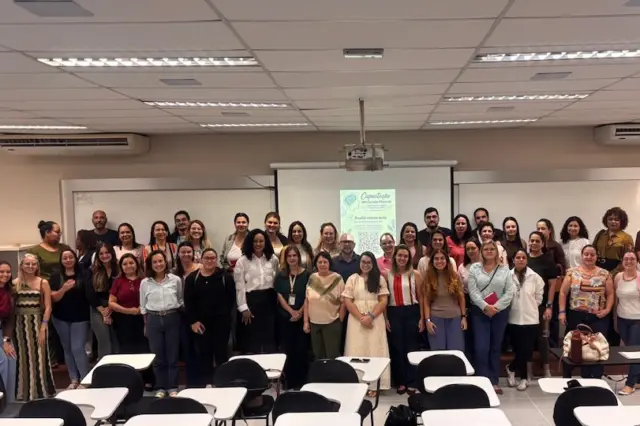Grupo de pessoas posando em sala de aula com tela de projeção e carteiras no primeiro plano