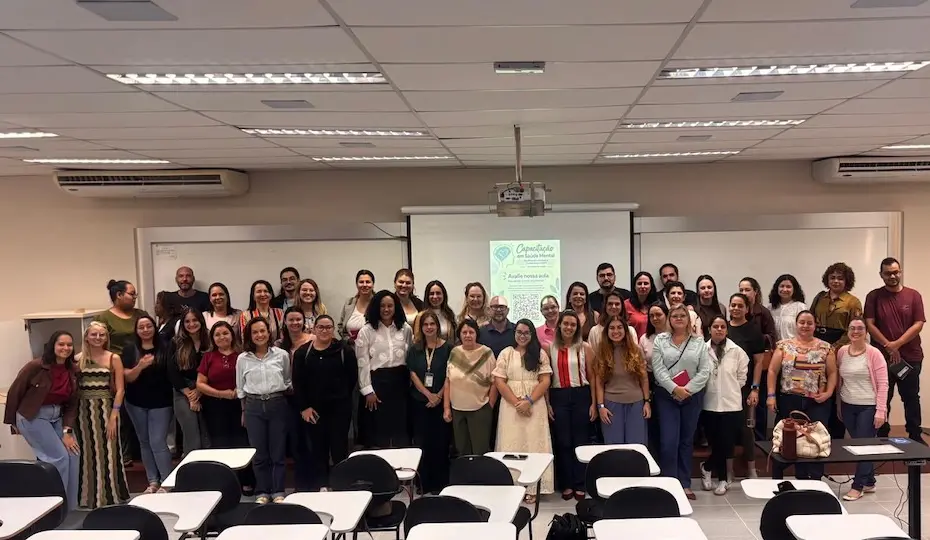 Grupo de pessoas posando em sala de aula com tela de projeção e carteiras no primeiro plano
