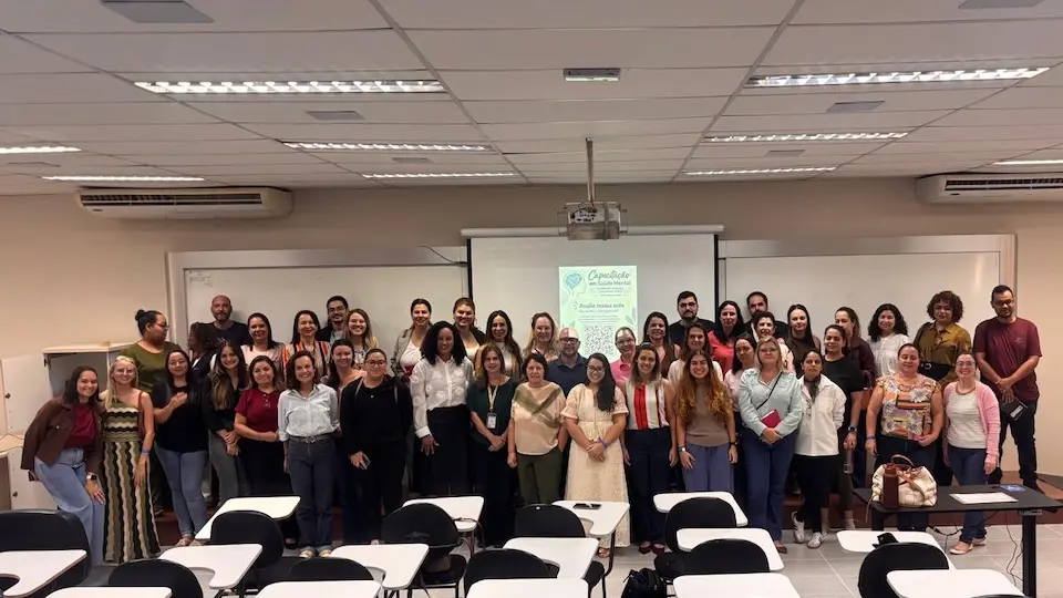 Grupo de pessoas posando em sala de aula com tela de projeção e carteiras no primeiro plano