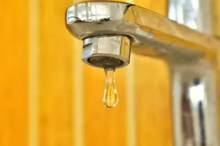 Torneira metálica com uma gota de água prestes a cair e fundo amarelado