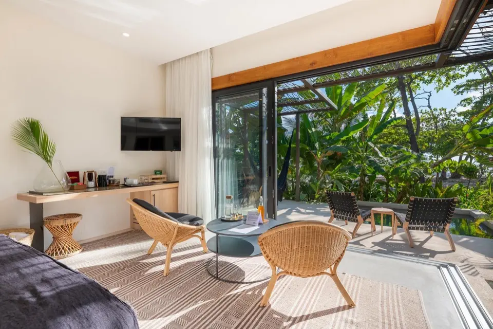 Quarto com portas de vidro deslizantes que se abrem para deck e vegetação tropical, cadeiras e mesa baixa