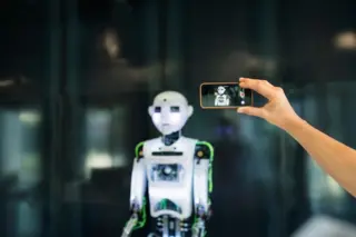Robô humanoide branco com detalhes verdes em exibição, e uma mão segurando um celular que registra o robô na tela.