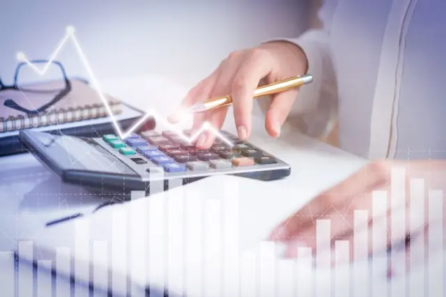 mão pressionando calculadora com caneta dourada na outra mão, bloco de notas e óculos ao fundo, com gráficos sobrepostos