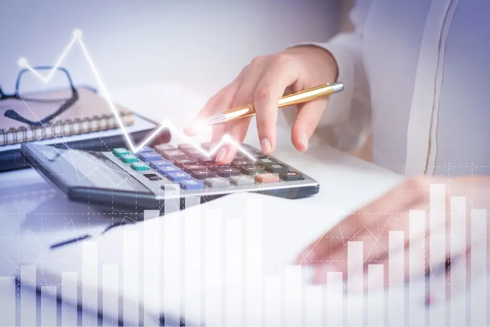 mão pressionando calculadora com caneta dourada na outra mão, bloco de notas e óculos ao fundo, com gráficos sobrepostos