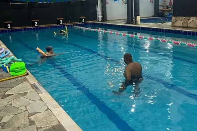 Piscina semiolímpica coberta com duas pessoas na água, um homem em profundidade média e outra pessoa com boia, e acessórios de hidroginástica na borda
