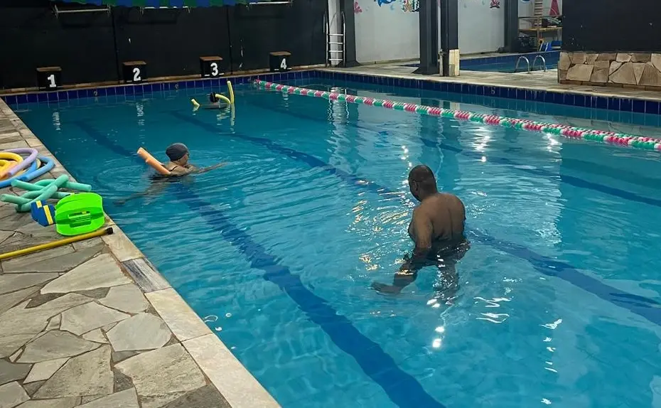 Piscina semiolímpica coberta com duas pessoas na água, um homem em profundidade média e outra pessoa com boia, e acessórios de hidroginástica na borda