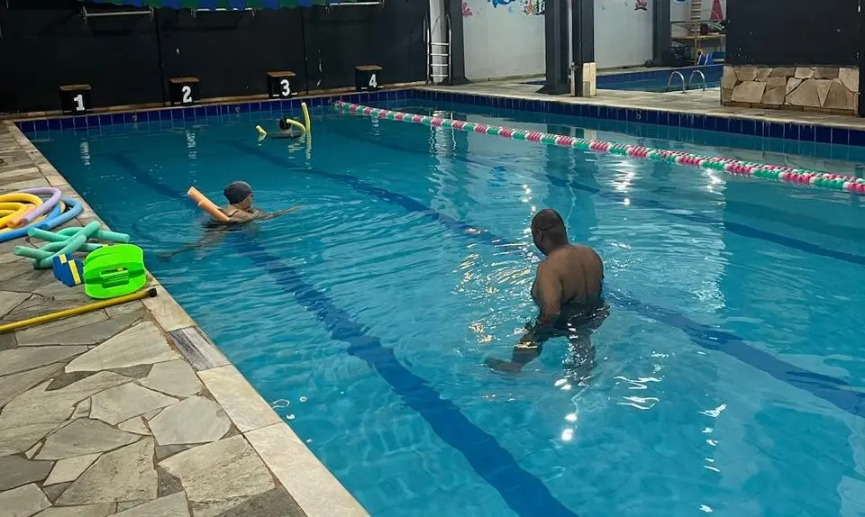 Piscina semiolímpica coberta com duas pessoas na água, um homem em profundidade média e outra pessoa com boia, e acessórios de hidroginástica na borda