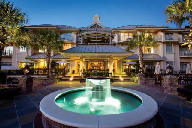 Fachada de hotel com fonte circular iluminada em primeiro plano, palmeiras e entrada com marquise ao anoitecer
