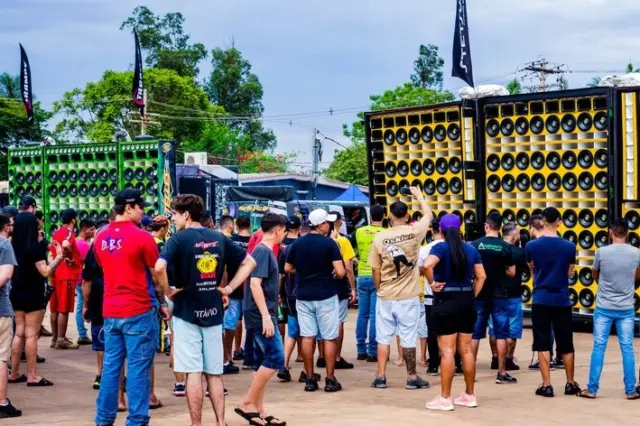 Pessoas reunidas em área externa observando caminhões e carretas equipadas com grandes painéis de alto‑falantes coloridos