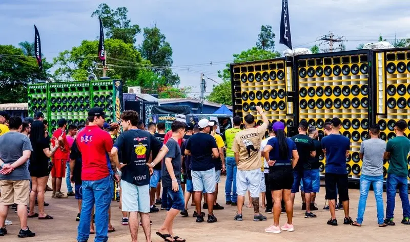 Pessoas reunidas em área externa observando caminhões e carretas equipadas com grandes painéis de alto‑falantes coloridos