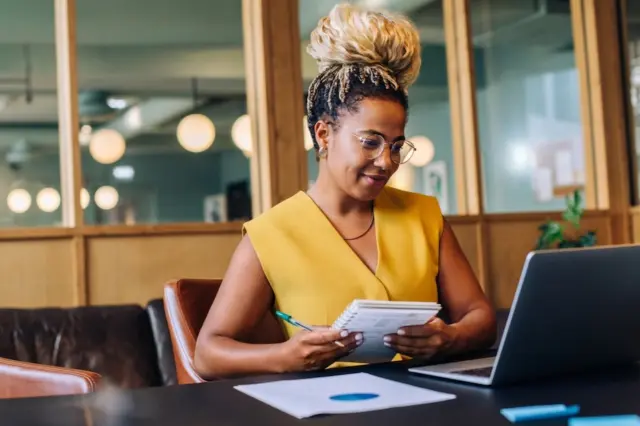 Mulher sentada em mesa de escritório usando óculos, segurando caderno e caneta diante de um laptop