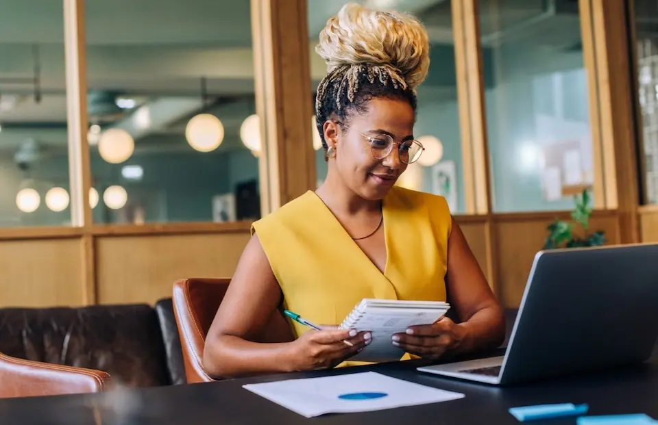 Mulher sentada em mesa de escritório usando óculos, segurando caderno e caneta diante de um laptop