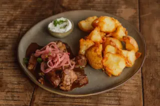 prato com pedaços de carne ao molho, cebolas em conserva rosadas, batatas douradas e um molhinho branco em recipiente pequeno, servido em prato rústico sobre mesa de madeira