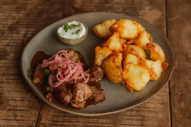 prato com pedaços de carne ao molho, cebolas em conserva rosadas, batatas douradas e um molhinho branco em recipiente pequeno, servido em prato rústico sobre mesa de madeira
