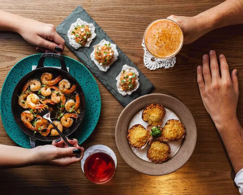 vista superior de mesa com frigideira de camarões, canapés e porção empanada, além de bebidas e mãos de duas pessoas