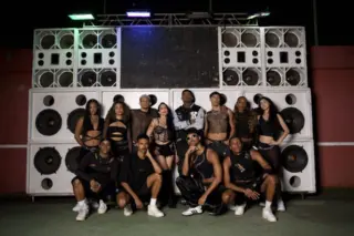 Grupo de dez pessoas vestidas em roupas pretas posando em frente a um grande paredão de alto-falantes