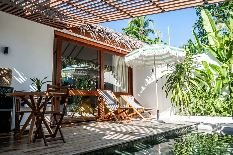 Deck de villa com cadeiras de madeira, espreguiçadeiras e guarda-sol ao lado de piscina e vegetação tropical