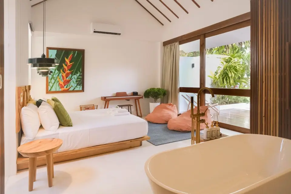 Quarto de villa com cama, banheira freestanding em primeiro plano e janelas para jardim