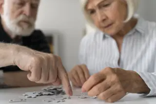 Duas pessoas idosas manuseando peças de quebra-cabeça sobre uma mesa; mãos em foco, rostos desfocados ao fundo