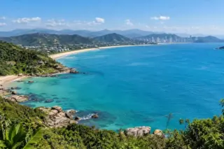 Vista panorâmica do litoral de Santa Catarina, com mar azul-turquesa, faixa de areia, morros verdes e prédios ao fundo.