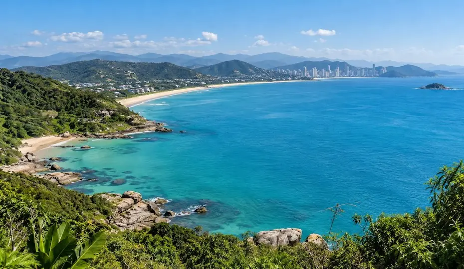 Vista panorâmica do litoral de Santa Catarina, com mar azul-turquesa, faixa de areia, morros verdes e prédios ao fundo.