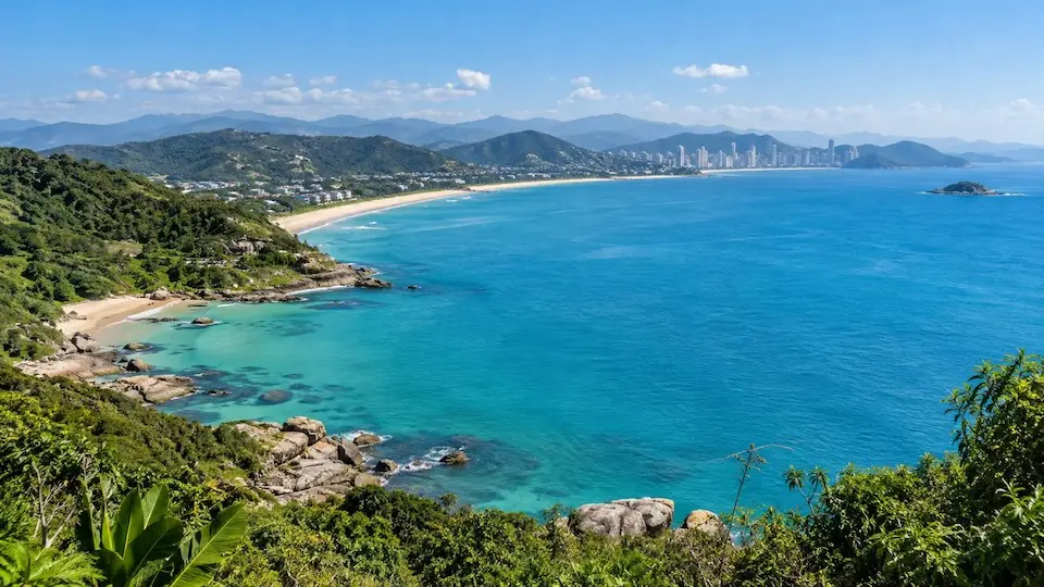Vista panorâmica do litoral de Santa Catarina, com mar azul-turquesa, faixa de areia, morros verdes e prédios ao fundo.