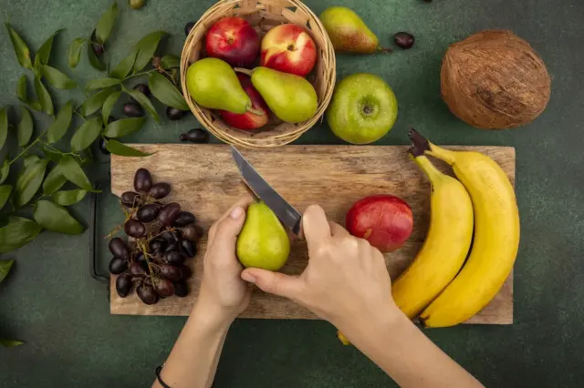 Mãos segurando uma pera e cortando com faca sobre tábua de madeira, cercada por bananas, cacho de uvas escuras, nectarinas, maçã verde e um coco