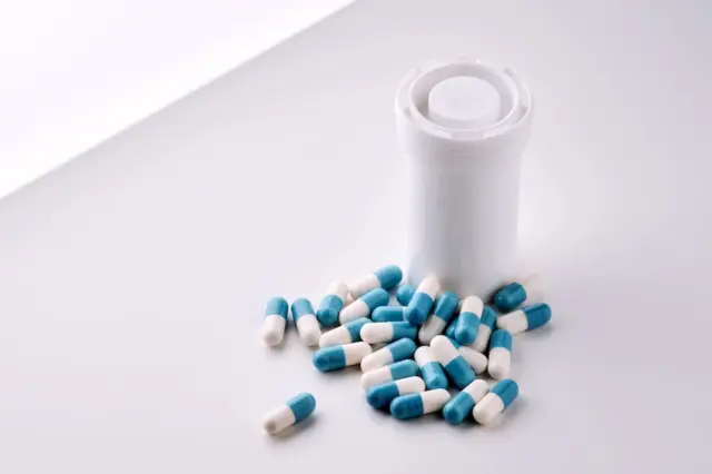 Frasco branco de medicamento ao lado de cápsulas azul e branca espalhadas sobre superfície clara